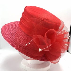 May include: A bright red hat with a woven straw texture on the brim and a smooth satin band around the crown. The hat is decorated with a large, layered red tulle flower accent featuring thin, delicate stems tipped with small white beads.