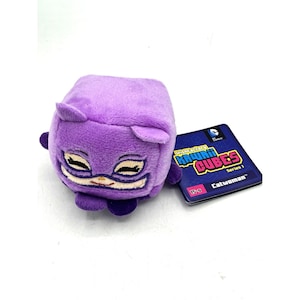 Op de afbeelding: Een paars pluche kubusvormig speelgoed in de vorm van Catwoman met kleine oortjes en geborduurde gelaatstrekken. Daarnaast ligt een rechthoekige verzamelkaart met het label Wish Factory Kawaii Cubes Series 1 Catwoman en een DC Comics logo.