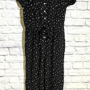 May include: A black jumpsuit featuring a small white and red floral pattern, short sleeves, a collared neckline, and a front button placket with a tie-front waist detail. The garment has a straight-leg silhouette and is displayed on a mannequin.