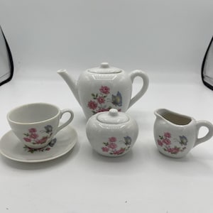 May include: A white ceramic tea set featuring a teapot, sugar bowl, creamer, teacup, and saucer. Each piece is decorated with pink roses and blue butterflies. The set is designed for a child's tea party.
