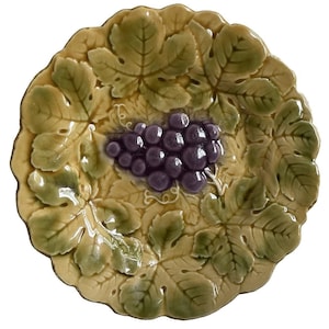 May include: A round ceramic plate featuring a raised, three-dimensional relief design of a cluster of purple grapes centered among textured, overlapping green and tan grape leaves. The plate has a scalloped edge and a glossy, glazed finish.