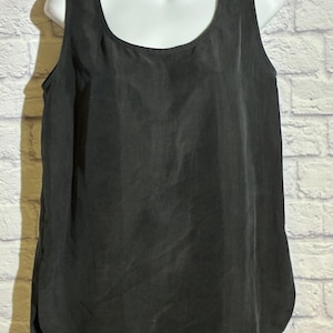 May include: A black sleeveless top with a scoop neckline. The top is made of a smooth, possibly silk-like material. The garment is displayed on a white mannequin torso against a white brick wall.