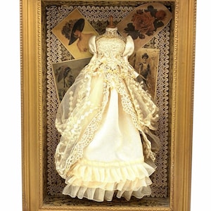 May include: A miniature dress displayed inside a rectangular gold frame. The dress is made of cream-colored fabric with lace overlays, a ruffled hem, and a high neckline. Behind the dress are vintage-style cards featuring illustrations of people.
