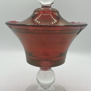 Könnte beinhalten: Eine rubinrote Glas-Kompottschale mit einem Sockel und Deckel aus klarem Glas. Die Kompottschale hat eine ausgestellte Schalenform und einen runden, klaren Glasknauf oben. Der Sockel ist aus klarem Glas mit rundem Fuß. Die Kompottschale wird wahrscheinlich zum Servieren von Desserts oder zum Ausstellen von Dekorationsartikeln verwendet.