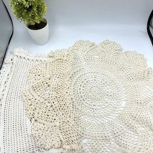 May include: Two off-white crochet doilies featuring intricate, openwork patterns. One doily is circular with a scalloped edge, while the other is rectangular with a grid-like center and a decorative border. Both pieces are crafted from fine thread.