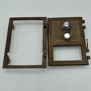 Pode incluir: Uma porta e moldura de caixa postal antiga na cor bronze. A porta apresenta dois botões prateados e um design decorativo de estrela. A moldura retangular tem uma porta com dobradiças e uma borda grega.