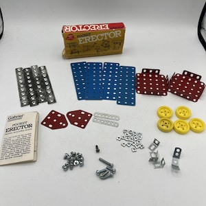 May include: A vintage Pocket Erector set with various metal and plastic construction pieces. Includes blue and red perforated plates, silver strips, yellow wheels, and a model-building guide. The box reads "Pocket Erector".