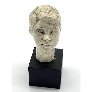 May include: A ceramic sculpture of a human head with a speckled off-white glaze, featuring defined facial features and a smooth hair texture. The head is mounted on a solid, matte black square base, creating a contrast between the light head and dark stand.