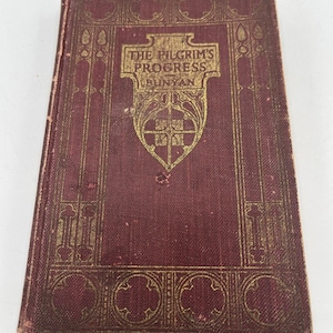 May include: A vintage book titled "The Pilgrim's Progress" by Bunyan. The cover is a deep burgundy color with gold embossed lettering and decorative borders. The book shows signs of wear, suggesting its age.