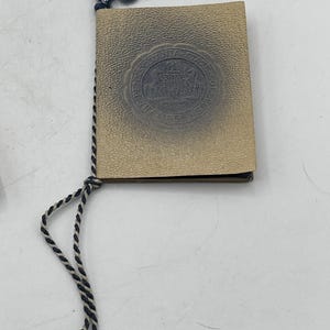 May include: A small, vintage book with a textured, tan cover. The cover features a circular emblem and text. A blue tassel and a black and white cord are attached to the book.