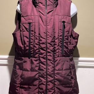 Puede incluir: Un chaleco acolchado burdeos con cuello alto y cremallera negra completa. El chaleco tiene dos bolsillos negros con cremallera y un diseño acolchado. El chaleco se exhibe en un maniquí blanco.