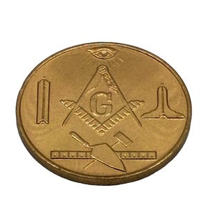 May include: A circular gold-toned metal coin featuring embossed Masonic symbols. The design includes a central square and compass with the letter G, an all-seeing eye above, a trowel, a level, a plumb rule, and a small hammer on the surface.