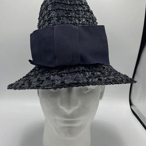 May include: A black straw hat with a wide brim and a large, dark bow. The hat is displayed on a white mannequin head. The hat's texture appears woven, and the bow is a solid, dark colour.