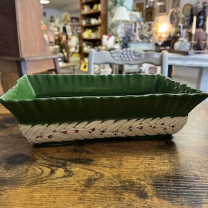 Puede incluir: Una jardinera rectangular de cerámica verde con un patrón decorativo de hojas blancas y rojas alrededor de la base. La jardinera tiene una superficie texturizada y está sobre una mesa de madera. La jardinera está vacía.