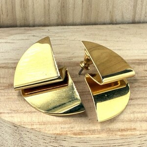 May include: A pair of gold-toned metal stud earrings featuring a geometric, semi-circular design with a folded, layered appearance. The earrings have a polished, reflective surface and include a standard post and clutch backing for secure attachment.