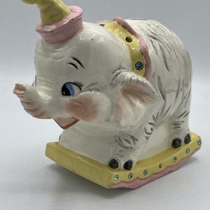 May include: A vintage ceramic elephant figurine, painted white with blue eyes, pink accents, and a yellow hat. The elephant is on a yellow base with pink trim and blue jewels. The figurine has a whimsical, circus-like design.