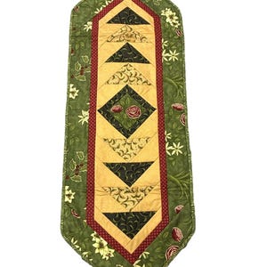 May include: A quilted table runner with a central design of gold, black, and red geometric shapes. The runner is bordered by a green floral pattern with white flowers and red berries. The runner is approximately 40 inches long.