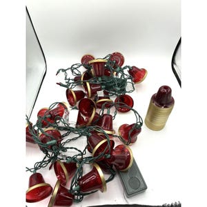 Peut inclure: Une guirlande lumineuse composée de cloches rouges translucides avec des bords dorés, reliées par un fil vert foncé. Une pile séparée de cloches rouges assorties avec des bords dorés est placée à droite de la guirlande lumineuse.
