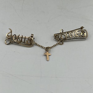 May include: A gold-coloured brooch set with the names "Jesus" and "Saves" in cursive script, linked by a chain with a small cross pendant. The brooch has a pin fastening.