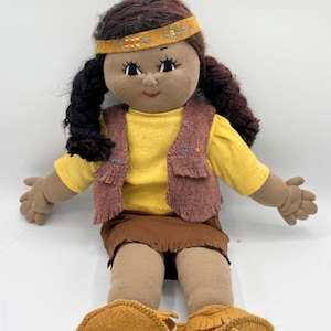 May include: A handmade doll with dark brown hair in braids, wearing a yellow shirt, a brown vest, and a brown skirt. The doll has a yellow headband with embroidered details and tan moccasins. The doll's skin is a medium brown color.