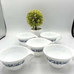 May include: A set of five white ceramic cups with a blue floral pattern around the exterior. Each cup features a wide, open bowl shape and a curved handle extending from the side. The cups are arranged in a group with a small green plant behind.