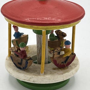 Puede incluir: Un carrusel de madera en miniatura con techo rojo, soportes amarillos y base blanca. El carrusel presenta pequeñas figuras montadas en caballos de madera. La base es verde y la parte superior tiene un botón dorado.