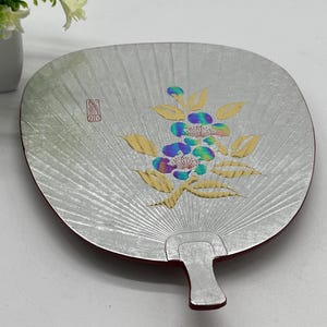 May include: A silver-colored, fan-shaped decorative plate with a red border and handle. The plate features a floral design with blue, purple, and yellow accents. A small red stamp is visible on the upper left side.