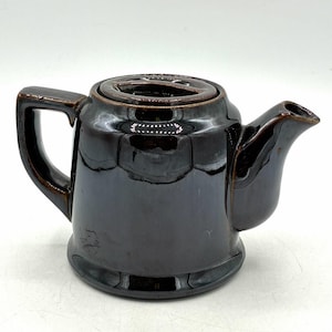 May include: A dark brown glazed ceramic teapot with a glossy finish. The teapot features a cylindrical body, a wide base, a curved handle on the left side, a short spout on the right side, and a circular lid with a recessed center handle.