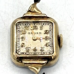May include: A vintage Gruen wristwatch with a gold-tone case and a square face. The watch has a cream-colored dial with black numerals and hands. The watch has a winding crown on the side and decorative gold accents.
