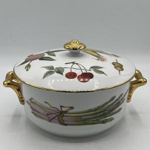 May include: White ceramic serving dish with a lid, featuring gold trim and handles. The dish is decorated with illustrations of asparagus, cherries, and other vegetables. The lid has a gold knob.