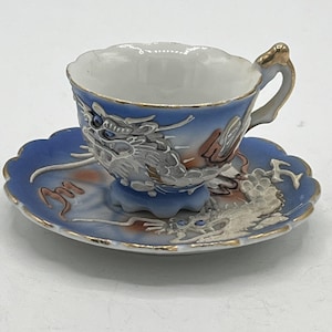 Op de afbeelding: Een kleine, sierlijke kop en schotel set. De kop en schotel zijn blauw met een wit drakenontwerp en gouden rand. De kop heeft een gebogen handvat met gouden accenten. De schotel heeft een geschulpte rand.
