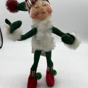 Puede incluir: Una caprichosa figura de elfo navideño con un gorro a rayas verdes y rojas, brazos y piernas de fieltro verde y zapatos de fieltro rojo. El elfo tiene un cuello y puños de piel blanca y una cara sonriente. Una pequeña etiqueta con el número 50 está adjunta.