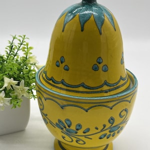May include: A yellow ceramic jar with a lid, adorned with teal blue floral patterns and accents. The jar has a rounded base and a matching lid with a decorative finial. The design features a combination of smooth and textured surfaces, creating a visually appealing piece.