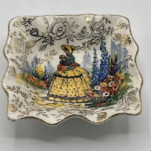 May include: A small, square, ceramic dish with a scalloped edge. The dish features a painted scene of a woman in a yellow dress in a garden, surrounded by flowers. Gold floral accents and trim adorn the white background.