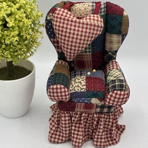 May include: A miniature armchair pincushion featuring a patchwork fabric design in shades of red, blue, green, and cream. The chair includes a heart-shaped pillow attached to the backrest, a ruffled skirt base, and two small sewing pins inserted.