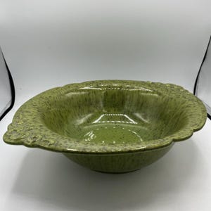 Puede incluir: Un cuenco de cerámica verde con un patrón moteado y un borde festoneado. El cuenco tiene una forma ancha y poco profunda y asas decorativas a cada lado. El interior es de color verde brillante.