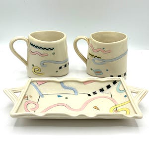 May include: Two off-white ceramic mugs and a matching rectangular tray with handles. The set features hand-painted abstract designs in pastel pink, light blue, yellow, and black, including squiggles, dots, and geometric shapes.