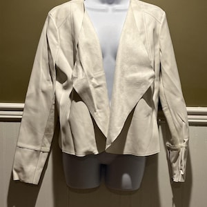 May include: A cream-colored jacket with a draped collar and long sleeves. The jacket is displayed on a white mannequin. The sleeves have a cuff detail. The jacket appears to be made of a soft, suede-like material.