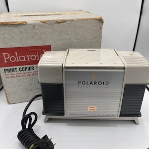 May include: A vintage Polaroid Print Copier Model 240, with a cream and black color scheme. The front features the Polaroid logo and the text "PRINT COPIER". The original box is visible in the background, with the Polaroid logo and product information.