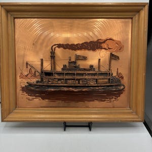 Könnte beinhalten: Ein rechteckiges Wandbild mit einem erhabenen Kupferrelief eines Raddampfers auf dem Wasser. Die Szene zeigt einen Schornstein mit dunklem Rauch, Flaggen und Bäume im Hintergrund. Das Kunstwerk ist in einen hellbraunen Holzrahmen gefasst.