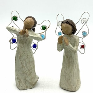May include: Two standing figurines with brown hair and cream-colored robes, each featuring wire-formed wings decorated with small, colorful glass beads in shades of green, blue, red, and white. The figures have smooth, featureless faces and hands held together.