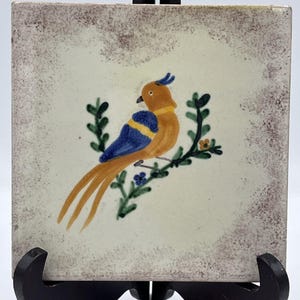 May include: Square ceramic tile featuring a hand-painted bird design. The bird has orange, blue, and yellow coloring, perched on a green branch with small blue flowers. The tile has a cream-colored background with a brown border. The tile is displayed on a black stand.