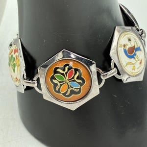 May include: A silver-toned link bracelet featuring multiple hexagonal charms. Each charm contains a circular enamel inlay with distinct colorful designs, including a central orange piece with a black floral motif and multicolored petals.