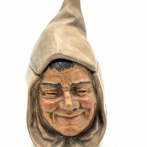 Op de afbeelding: Een keramieken beeldje van een hoofd met een puntige kap. Het gezicht heeft gedetailleerde rimpels, een subtiele glimlach en donker geschilderde wenkbrauwen en haar. De kap heeft een matte bruine tint en het gezicht is bruin getint.