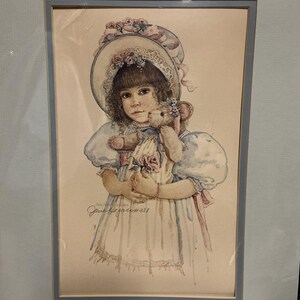 Könnte beinhalten: Aquarelldruck eines jungen Mädchens in einem weißen Kleid und Hut, das einen Teddybären hält. Das Mädchen hat braune Haare und trägt ein hellblaues Kleid mit Puffärmeln. Der Druck ist in einem hellblauen Rahmen gerahmt.