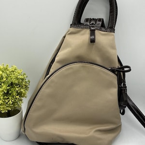 May include: A beige nylon sling backpack featuring a triangular silhouette, dark brown leather-like trim, a top handle, and a semi-circular front zippered pocket. The bag includes adjustable straps with silver-toned metal hardware and zipper pulls.