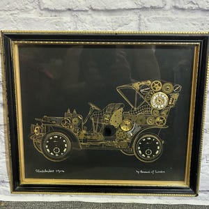 Op de afbeelding: Een ingelijst kunstwerk met het silhouet van een oldtimer, opgebouwd uit goudkleurige tandwielen en horlogeonderdelen op een zwarte achtergrond. De tekst Studebaker 1904 en by Ammon of London staat in het wit onderaan de zwarte passe-partout.