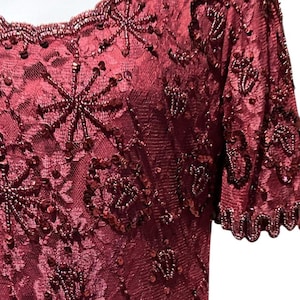 May include: Burgundy-colored formal dress with intricate bead and sequin embellishments. The dress features a scoop neckline and short sleeves, with detailed floral and starburst patterns. The fabric appears to be a sheer lace overlay.