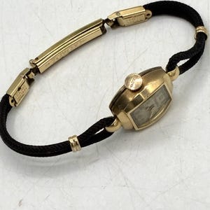 May include: A vintage Bulova wristwatch featuring a gold-tone oval case with a matching crown and a light-colored dial. The watch is attached to a black cord strap accented with gold-tone metal links and a rectangular clasp engraved with U.S.A. PAT.