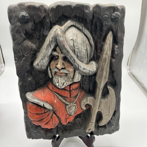 May include: A rectangular, carved wall plaque depicting a relief of a man in a hat and red tunic, holding a weapon. The background is dark with a wood-grain texture. The man has a white beard and a pendant.